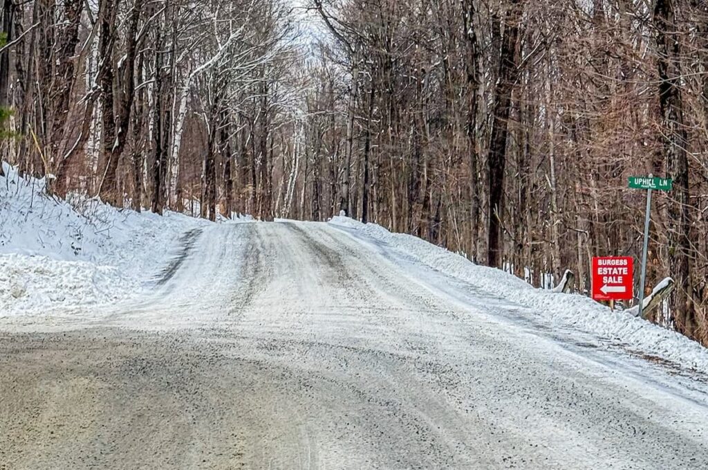 Snowy Vermont road in winter with a red sign and arrow that says Burgess Estate Sale.