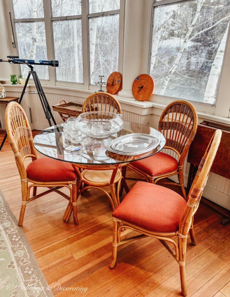 Wicker chairs with orange cushions around glass table and telescope in Vermont luxury estate sale home with birch trees out sunroom windows.