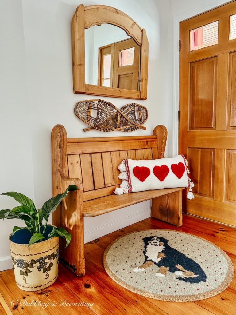 Church pew bench entryway with Bernese Mountain Dog rug, blueberry bucket with plant, wall mirror, snowshoes and Valentine's Day pillow.