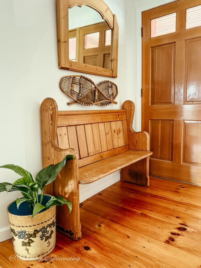 Church pew bench entryway with wood flooring, overhead mirror on wall with snow shoes and a blueberry bucket with green plant.