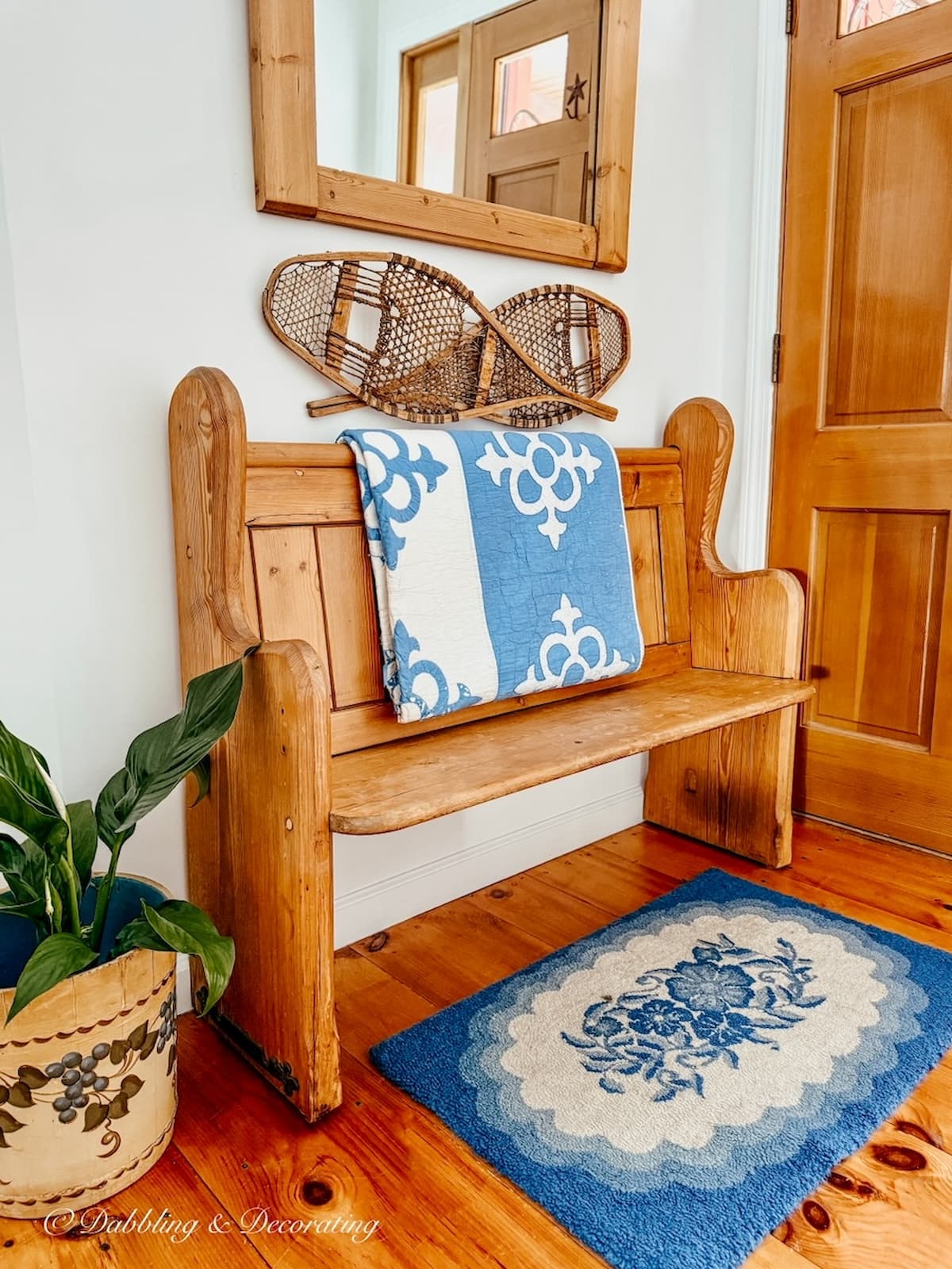 Vintage Pine church pew bench entryway decorated with vintage blue and white quilt, blue hooked rug, snow shoes, mirror and bucket with plant on floor.