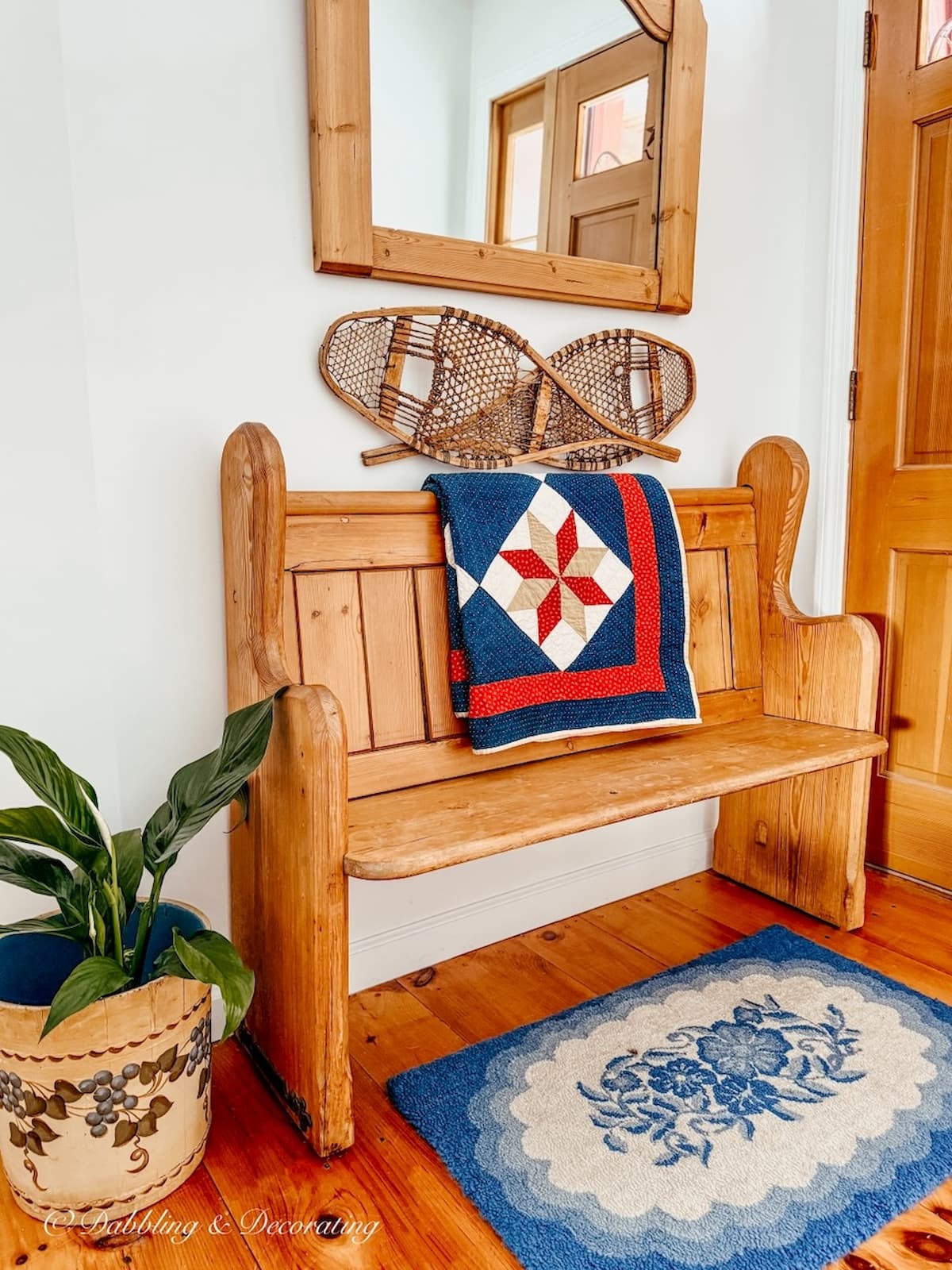 Vintage church pew bench entryway decorated with blue rug, blue and red quilt, snowshoes, mirror and vintage wooden blueberry bucket on floor with plant.