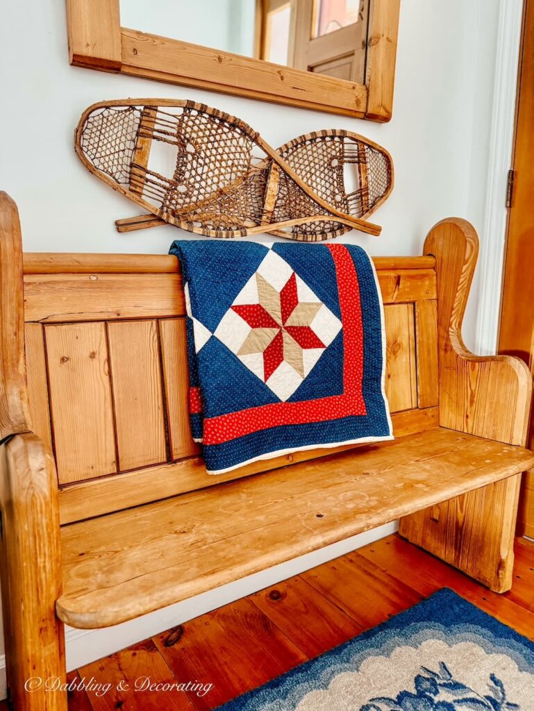 Church pew bench entryway decorated with red, white, and blue quilt, blue hooked rugs, snow shoes and wall mounted mirror.