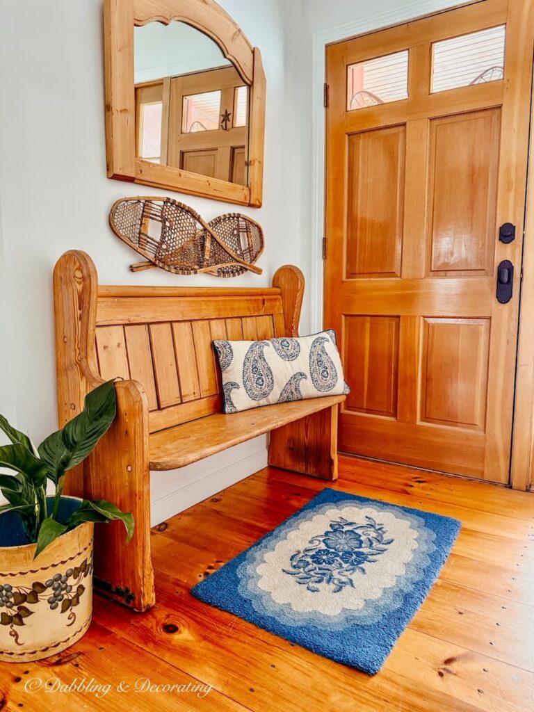 Church pew bench entryway decorated with a blue hooked rug, white and blue paisley pillow, snowshoes and wall mirror with vintage bucket with plant on floor.