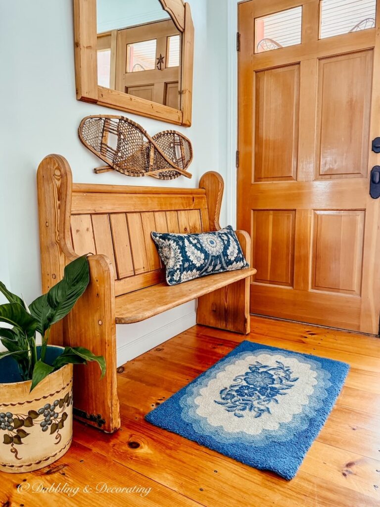 Church pew bench entryway decorated with a blue hooked rug, blue paisley pillow, snowshoes and wall mirror with vintage bucket with plant on floor.
