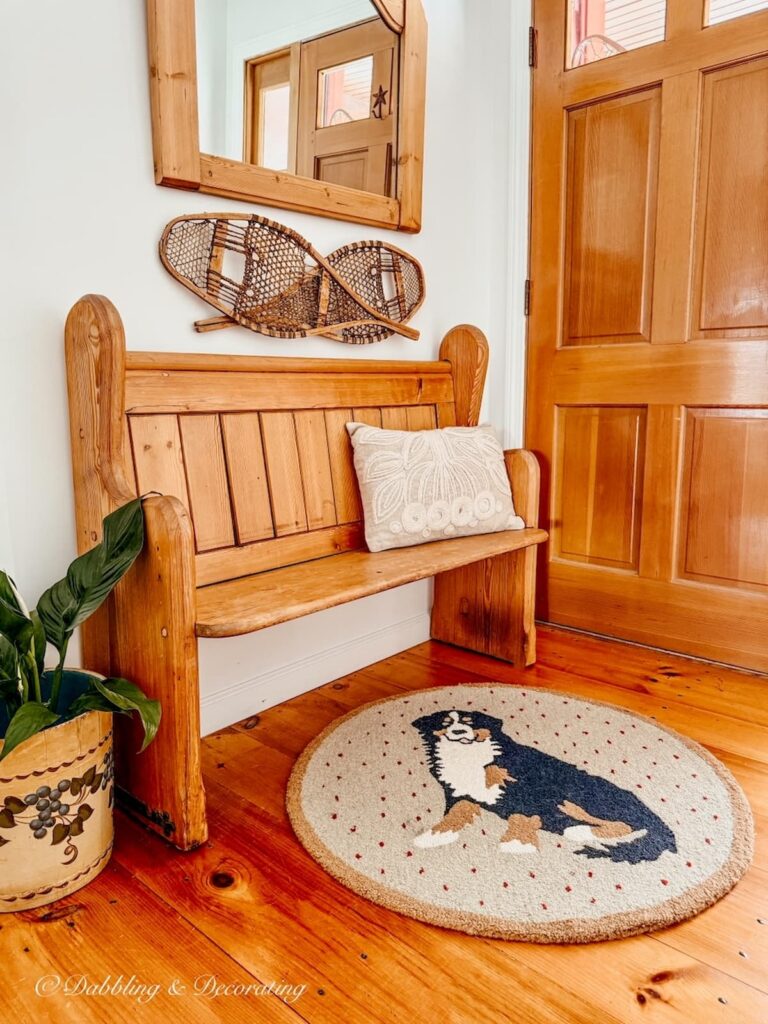 Vintage pine church pew bench entryway decorated with Berner rug, snow shoes, mirror and blueberry bucket with plant on floor.