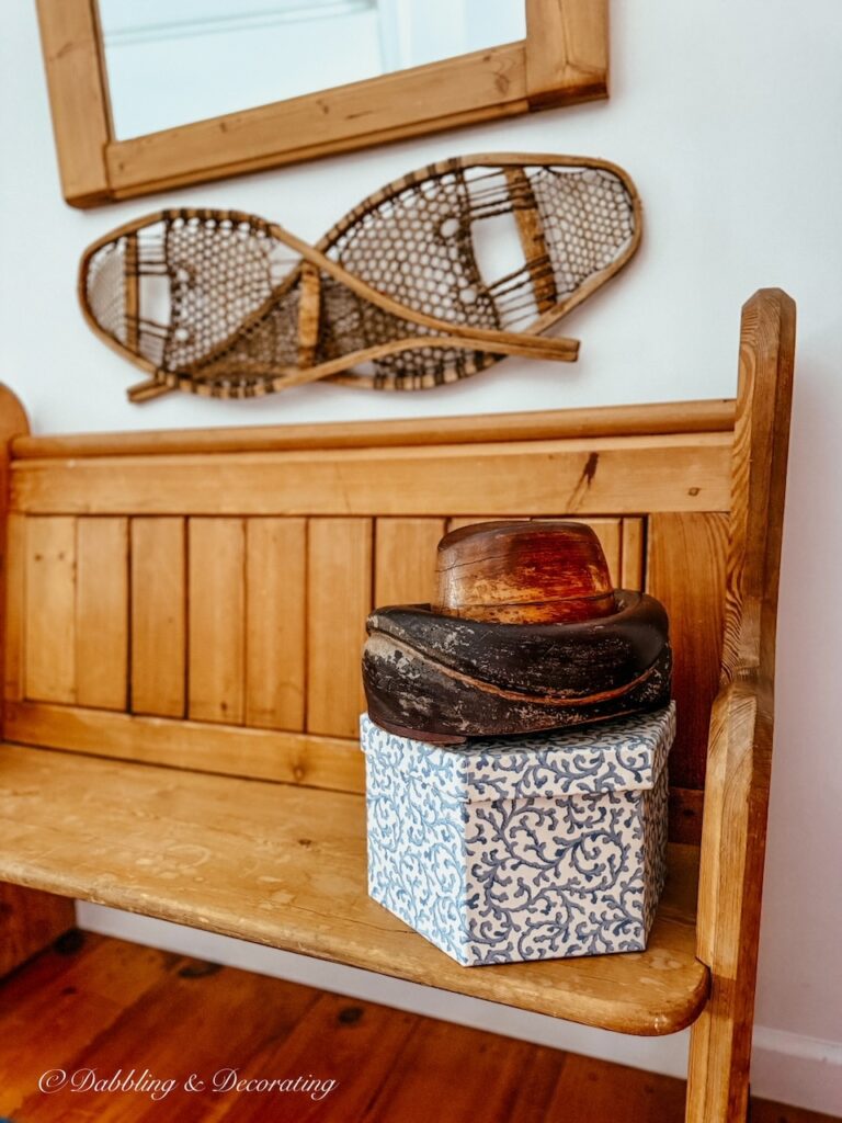 Antique hat mold on top of a blue and white hat box on vintage church pew bench entryway with snowshoes on wall.