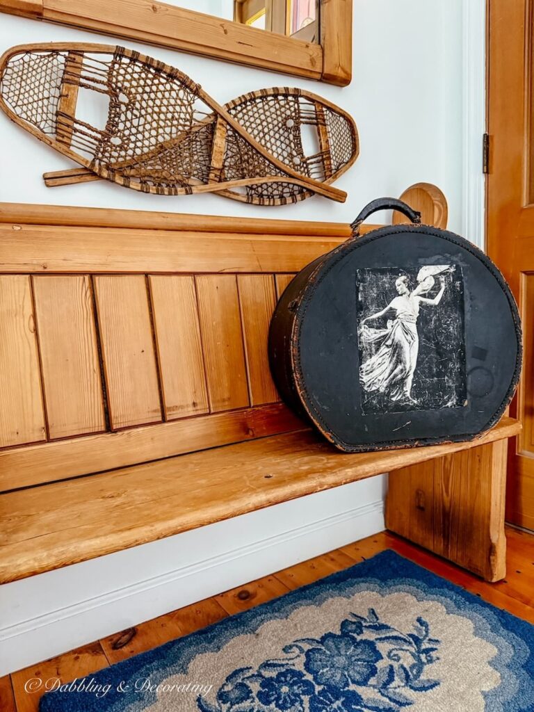 Vintage church pew bench entryway decorated with blue rug, antique black round suitcase and snow shoes on wall.