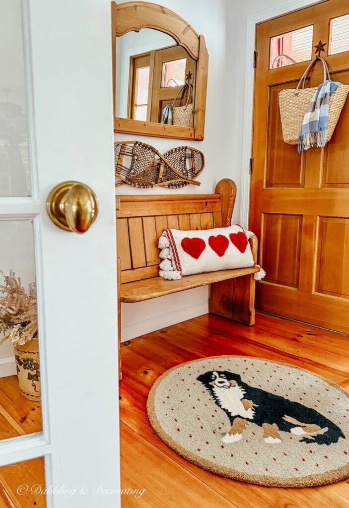 Church pew bench entryway decorated with Valentine's Day pillow, Dog rug, snowshoes and wall mirror and basket with scarf on front door.