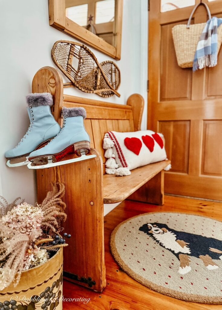 Vintage church pew bench entryway decorated with wall mirror, snowshoes, bucket of dried flowers, Valentine's Day pillow, dog hooked rug and blue vintage ice skates with basket and blue scarf on door.