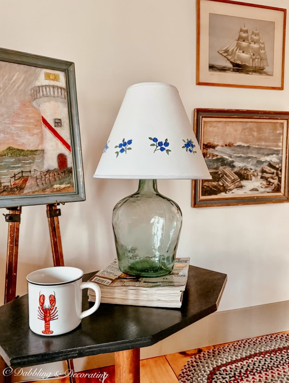 Coastal home vignette with vintage glass lamp and coastal art on wall and easel with a lobster mug in Maine.