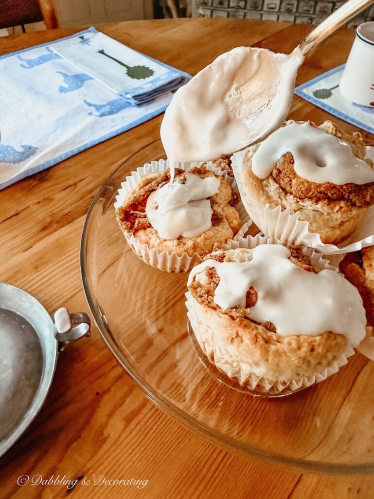 Cinnamon roll buns on a glass cakes stand on table with spoon pouring frosting on them. A Sunday home moment.