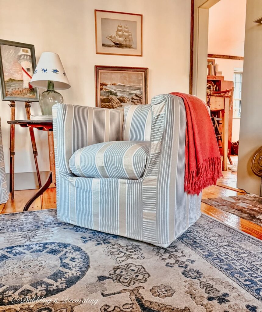 Living room slipcovered chair with pink blanket next to fireplace with vintage side table and wall art in coastal Maine home for a Sunday home decor vibe.
