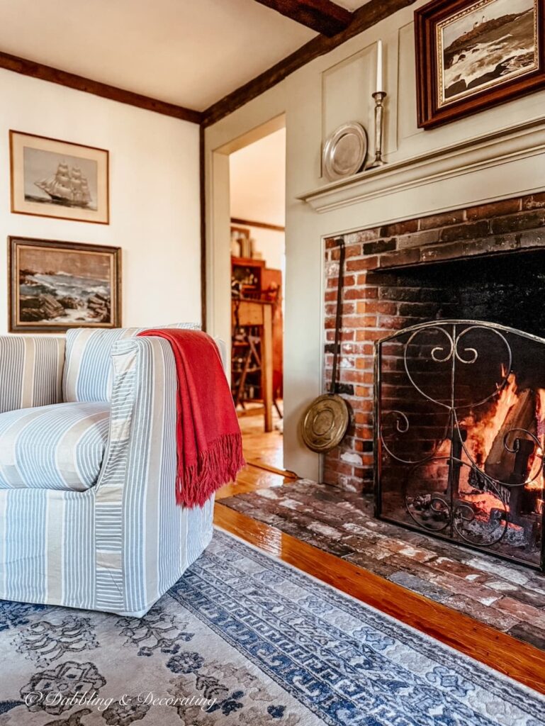 Cozy Sunday home vibe in colonial styled living room with chair in front of fireplace with a pink throw and fire in the fireplace.