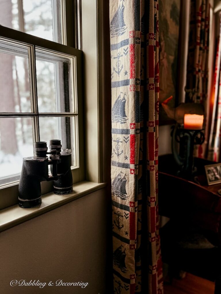 Vintage binoculars resting on a windowsill during a snowfall next to retro coastal drapes with lit candle for a Sunday home snow day vibe.