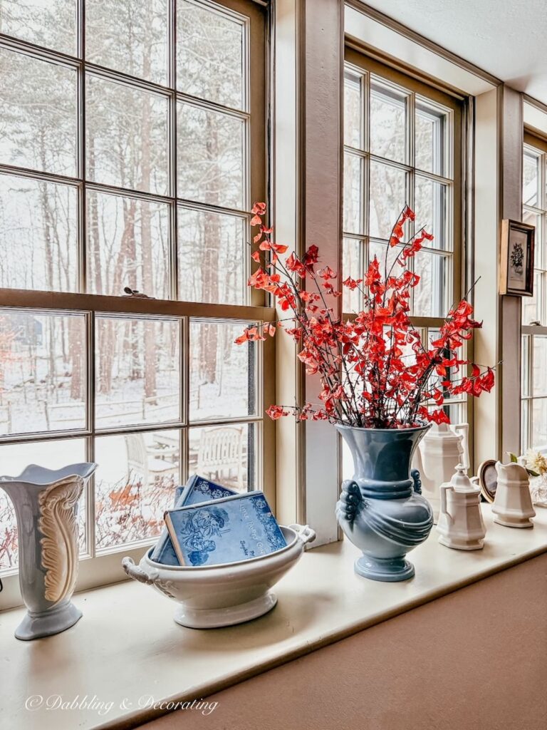 Long windowsill decorated with antiques and blue vase with red stemmed leaves and snow just out the window for a Sunday home vibe.