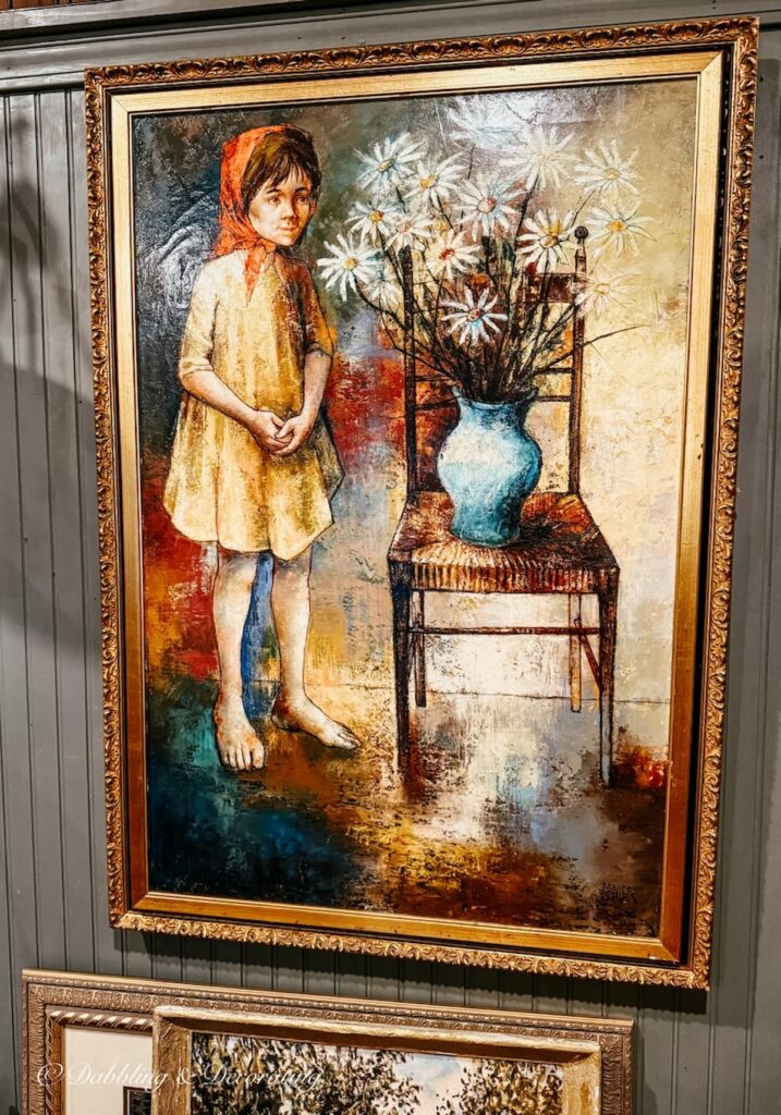 Antique painting on wall with gilded gold frame with small child looking onto a vase filled with flowers on antique chair.