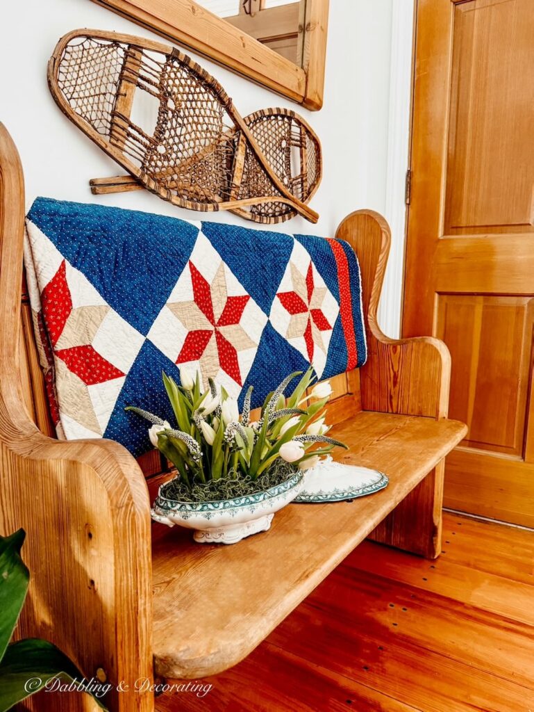 Vintage church pew in entryway with red, white, and blue quilt and vintage soup tureen planter ideas with spring flowers.