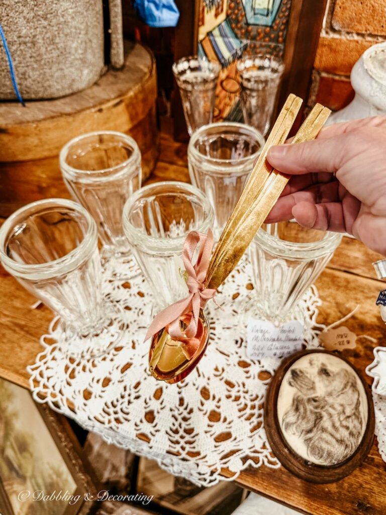 Set of dessert golden spoons in hand in front of authentic vintage soda fountain glasses at Waverly Square Antiques in Saratoga, NY.