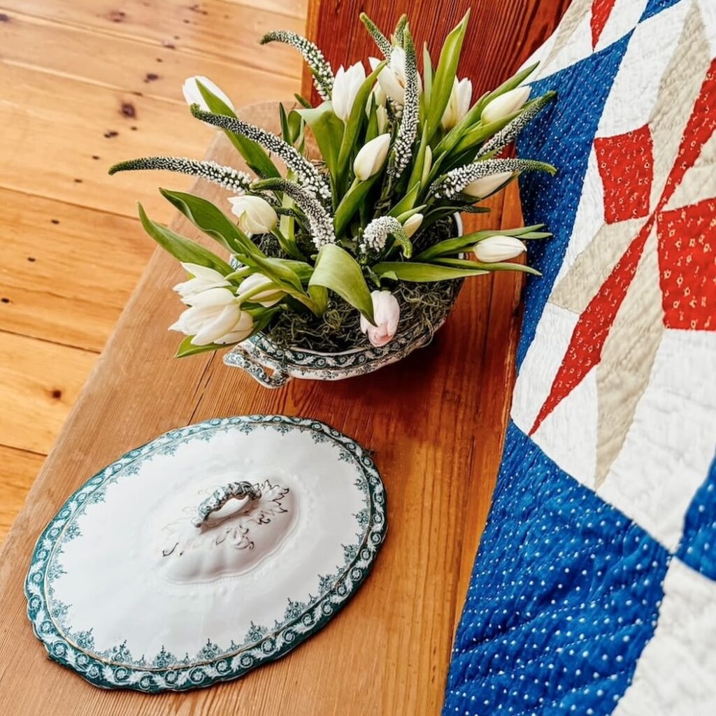 Vintage soup tureen displayed as a flower planter with white tulips on vintage church pew in entryway with red, white, and blue quilt.