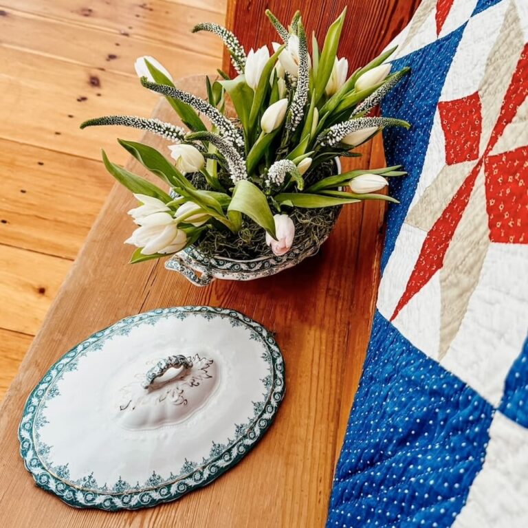 Vintage soup tureen displayed as a flower planter with white tulips on vintage church pew in entryway with red, white, and blue quilt.