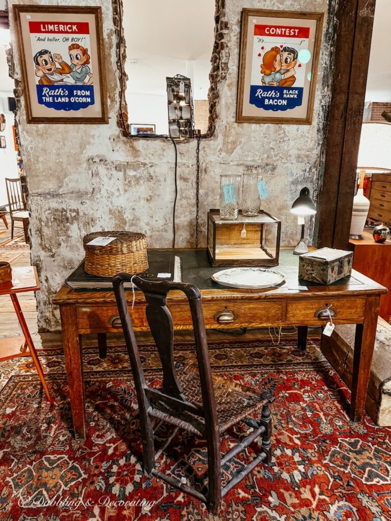 Antique booth styled with a red vintage turkish rug, antique desk and chair against brick wall with paintings.