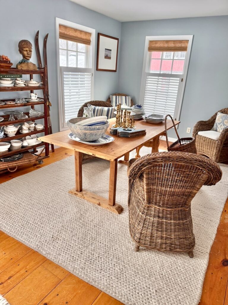 Corner room in an open floor plan before Scullery-Inspired Kitchen Workroom transformation with pine table, wicker chairs, white rug and vintage cobbler rack with antique finds.