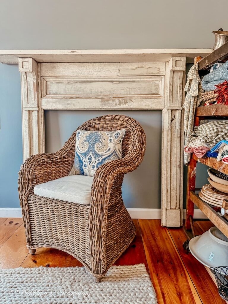 Room in home with blue walls and a vintage faux white mantel with wicker chair and cobbler rack rearranging furniture for a scullery-inspired kitchen workroom.