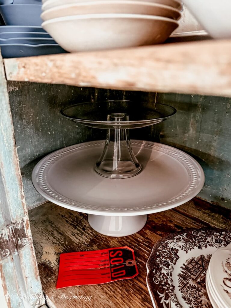 Inside view of sneak peak of an antique hutch in a soon to be scullery-inspired kitchen workroom with cake stands and vintage dishes and a sold tag.