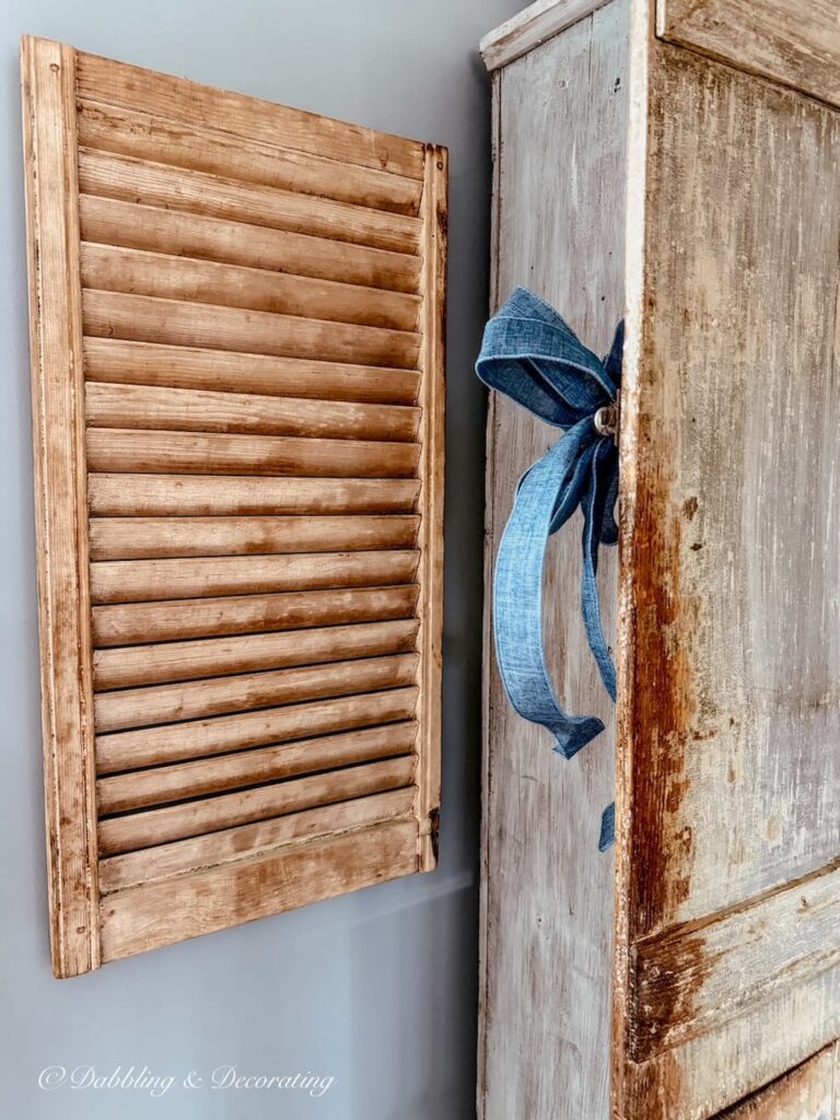 Vintage wooden shutters hanging on a blue wall next to an antique cupboard with a blue bow.