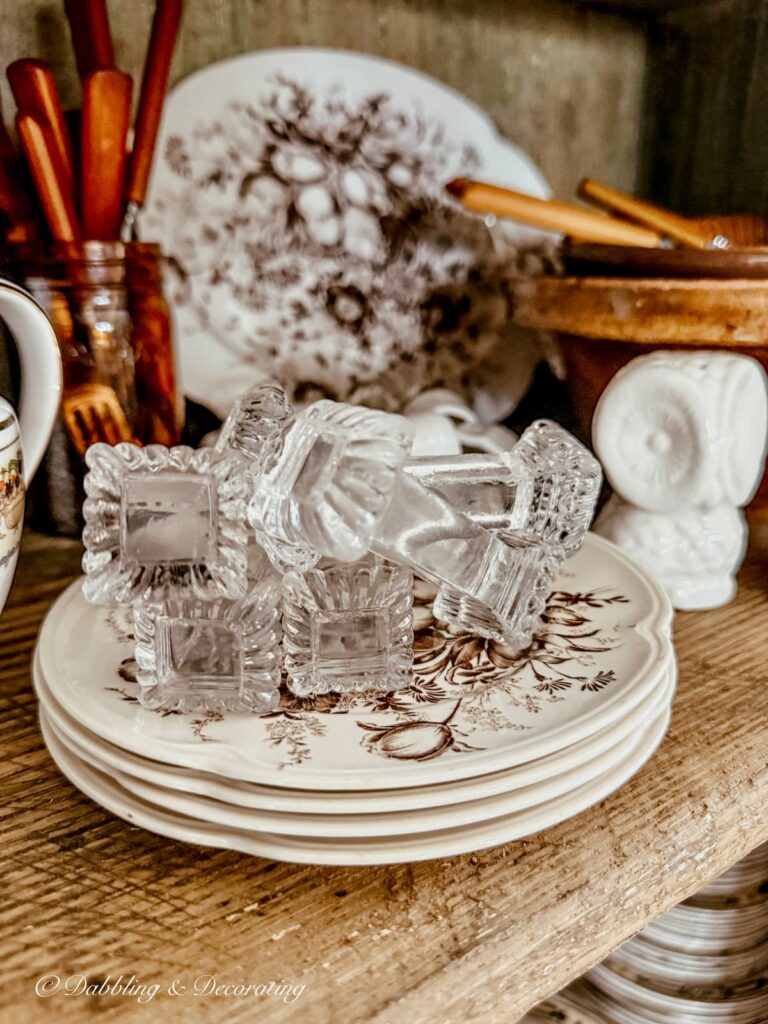 Stack of small brown and white transferware with a collection of crystal knife rests on antique cabinet shelf.