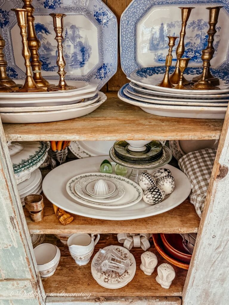 Vintage dishes, platters, candlesticks stacked and layered for antique cabinet ideas in scullery-inspired kitchen workroom.