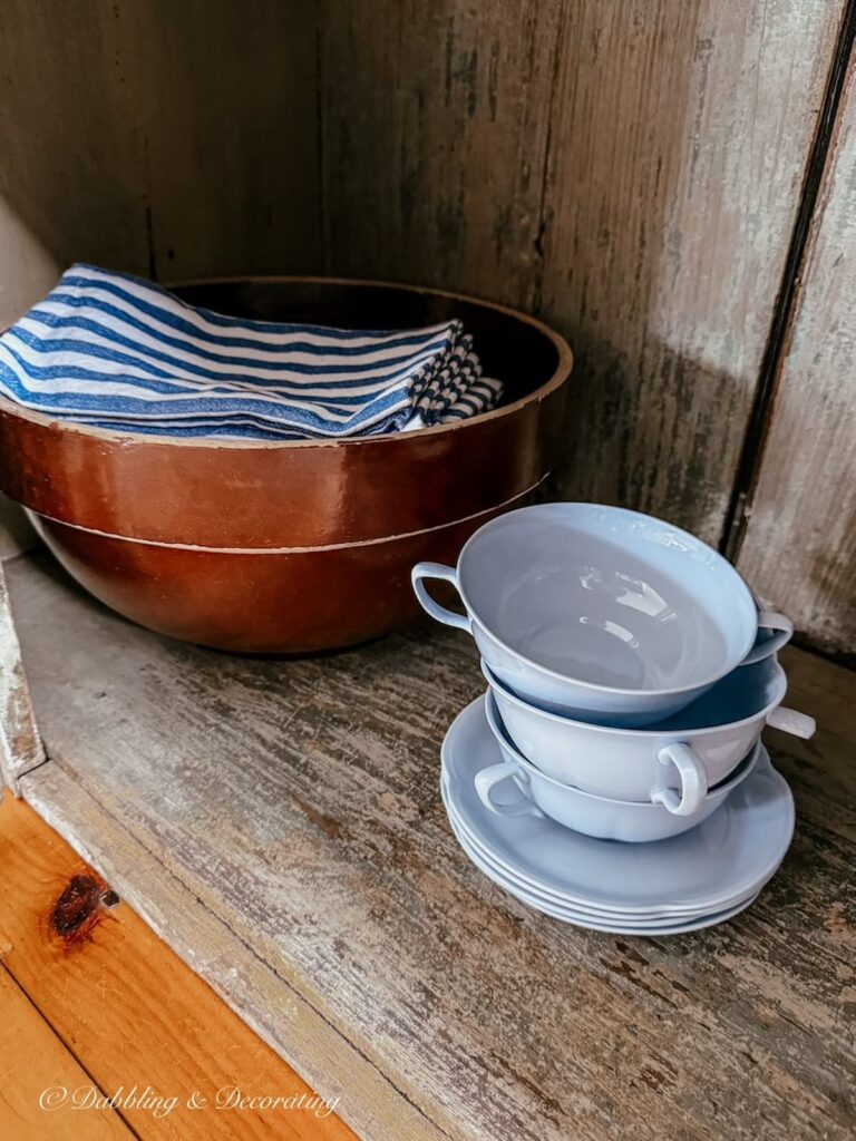 Inside an antique cupboard sits a large brown stoneware vintage bowl with blue striped napkins and a stack of blue vintage cream soup bowls and saucer for vintage decor storage ideas.