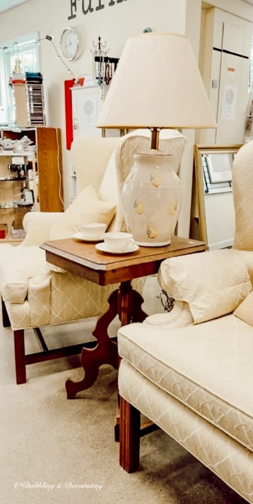 Vintage wooden table between two upholstered chairs with lamp in thrift store.