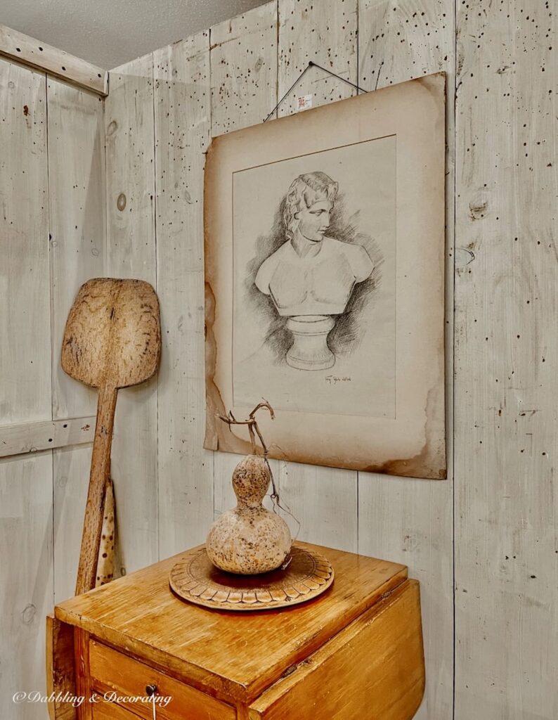 Simple Neutral Vintage booth display with wooden antique table, neutral drawing on wall, bread paddle leaning in corner and fall ghourd on table.