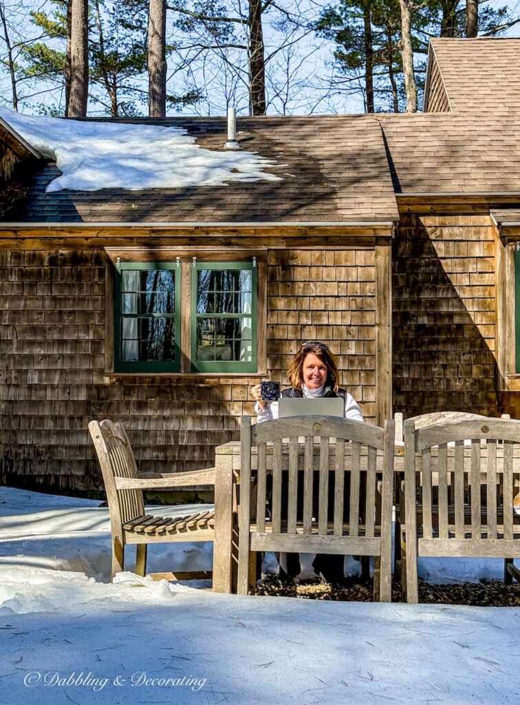 Ann, vintage home decor blogger sitting at outdoor teak table during mud season with snow at home in Maine.