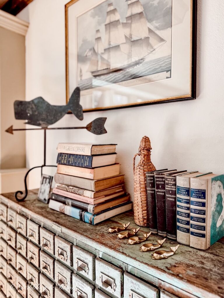 Antique apothecary cupboard styled with vintage books, a wrought iron whale weathervane and a vintage demijohn as a bookend in an antique Maine home.