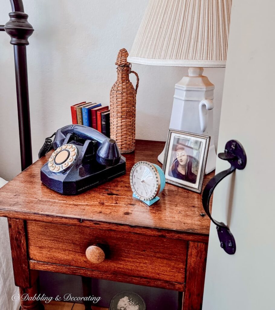 Antique wooden side table in bedroom with old telephone, vintage demijohn bottle, alarm clock in eclectic home decor.