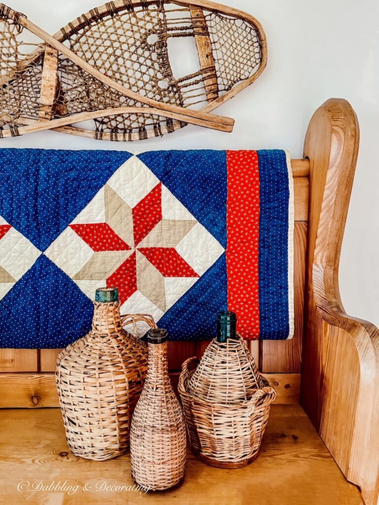 Three vintage demijohns on a vintage pine church pew with a draped blue and red quilt and snowshoes on wall in entryway in eclectic home decor ideas.