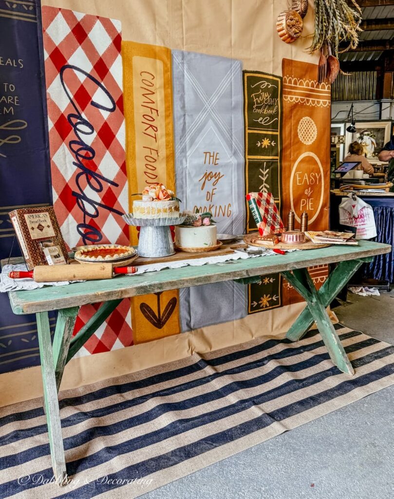 Vintage farmhouse green table with vintage kitchen items like cookbooks, runner, rolling pins, faux bakery on striped rub runner at Vintage Market Days.