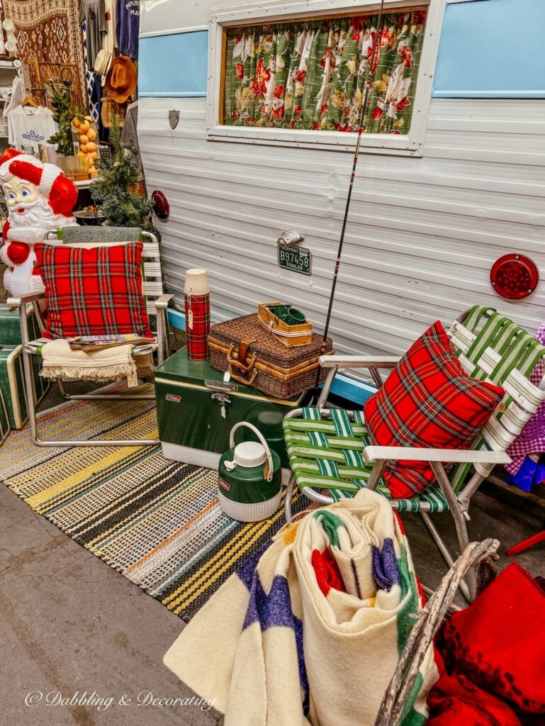 Vintage retro camping gear styled for sitting.  Coleman coolers, blankets, rug, plaid pillows and retro green chairs next to vintage camper at Vintage Market Days.