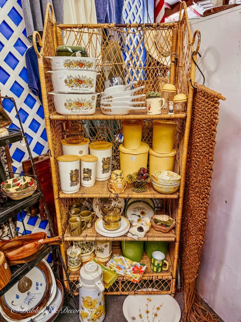 Wicker shelving in vintage booth styled with retro orange, yellow, and green tupperware, corningware, dishes, and more.