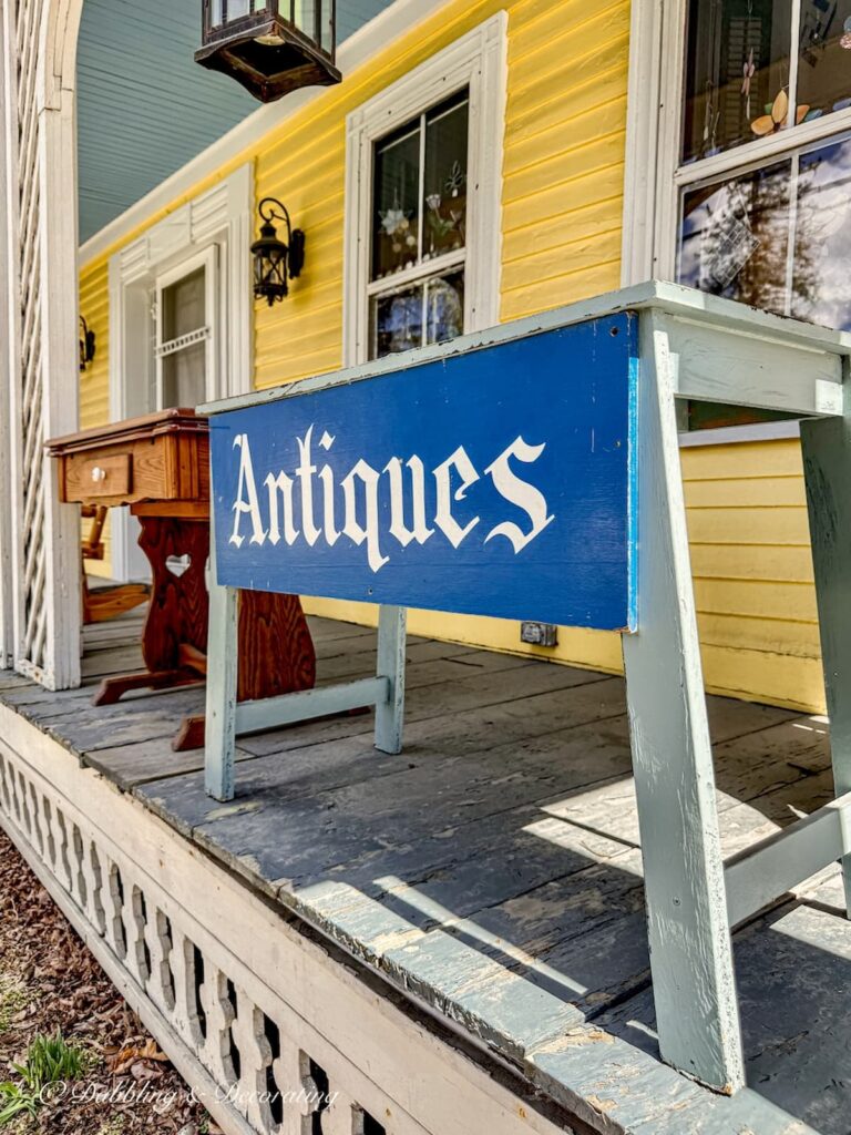 Antiques blue sign on porch of old yellow farmhouse in New England with antique furnishings. Solo antiquing road trip in Vermont spring morning
