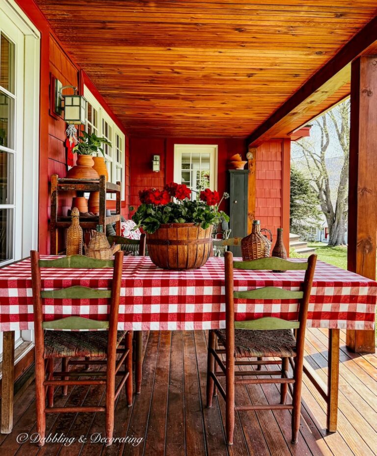 Covered porch table with red gingham tablecloth, vintage demijohns and a round crate filled with red geraniums for spring.