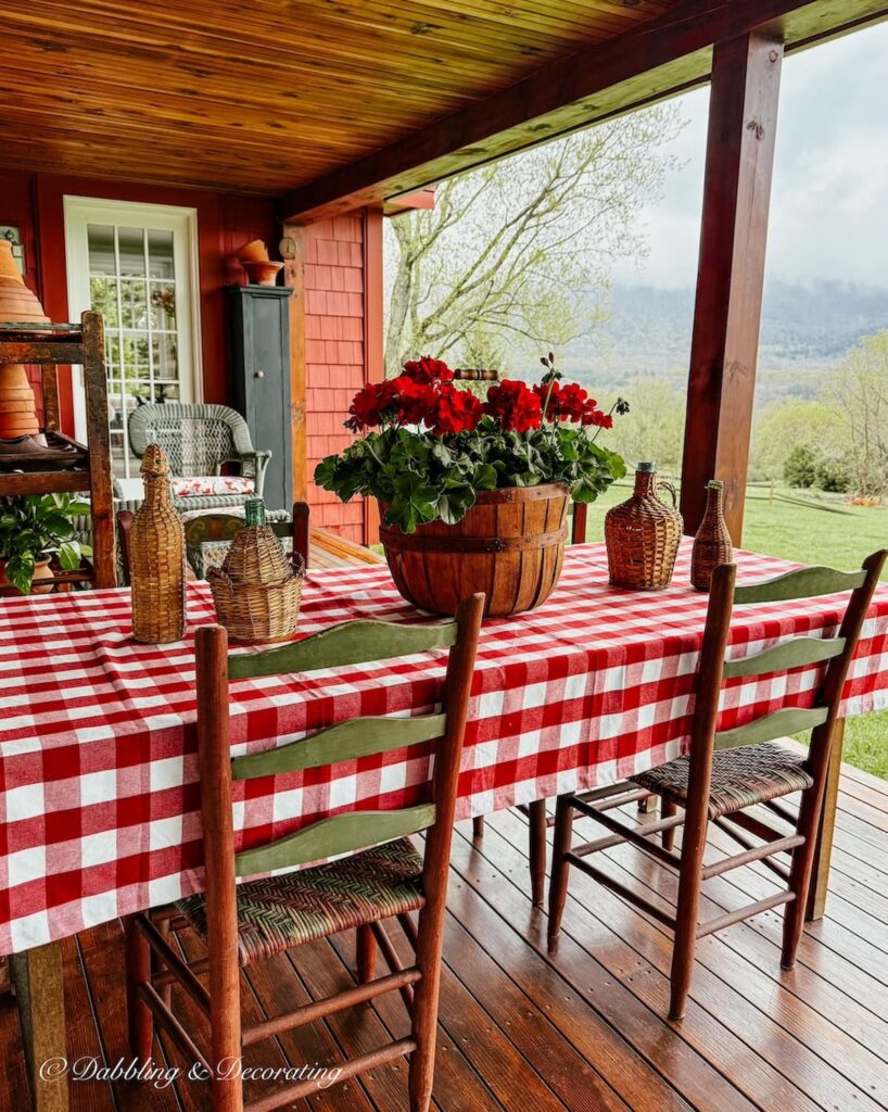Covered porch table with red gingham tablecloth, vintage demijohns and a round crate filled with red geraniums on a rainy mountain day.