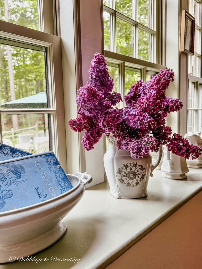 Lilac arrangement in brown and white transferware pitcher on large windowsill with ironstone tureen filled with blue books.