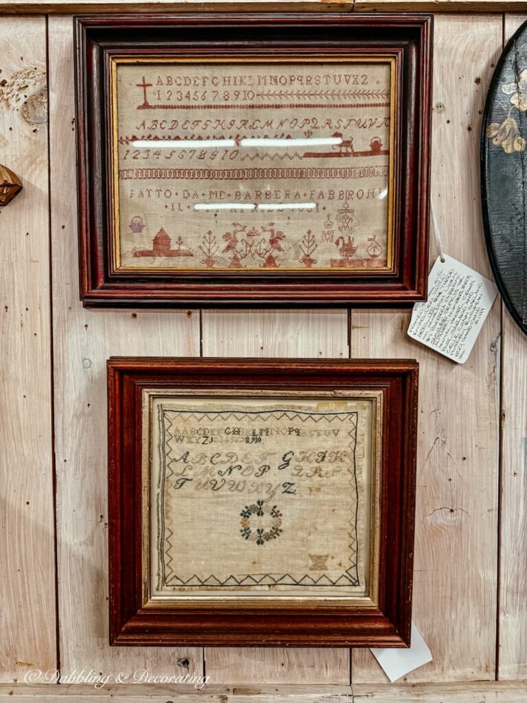 Needlepoint pictures in antique wooden frames on wall in vintage booth at antique store for Americana decor ideas.