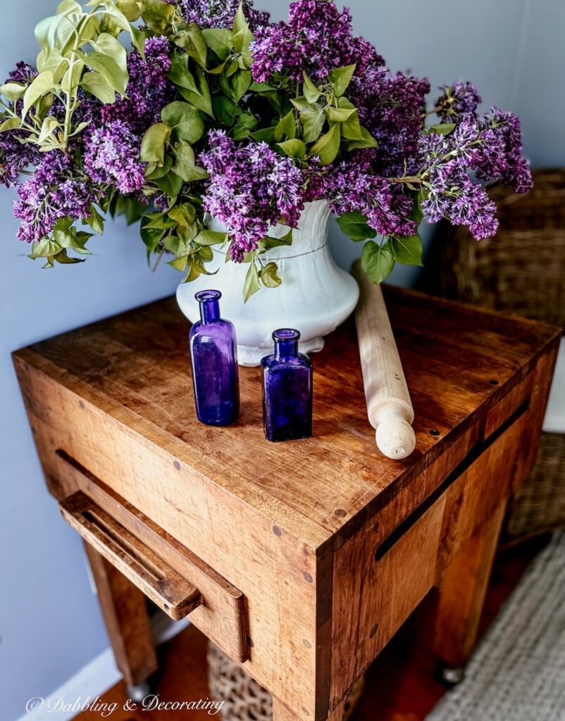 Vintage butcher block styled with spring lilacs in old white grub pot with amethyst bottles, and rolling in in scullery corner.