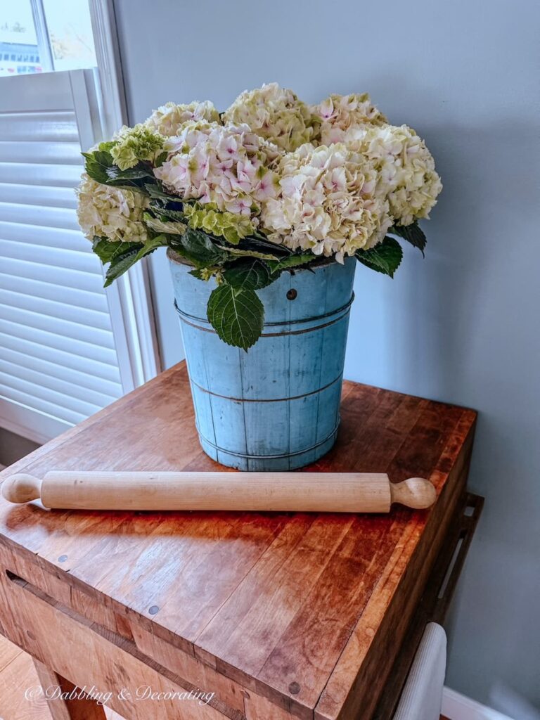 An antique blue ice cream bucket filled with white hydrangeas on a vintage butcher block table with an oversized rolling pin in kitchen scullery.