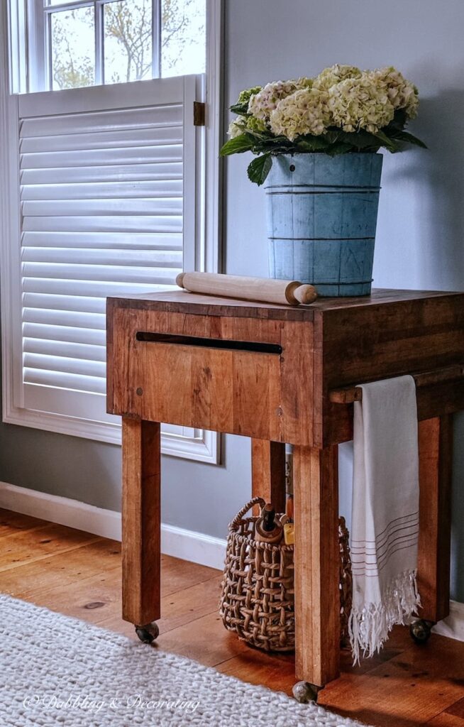 Vintage butcher block styled in scullery space with a blue bucket filled with hydrangeas and a basket of old rolling pins underneath.